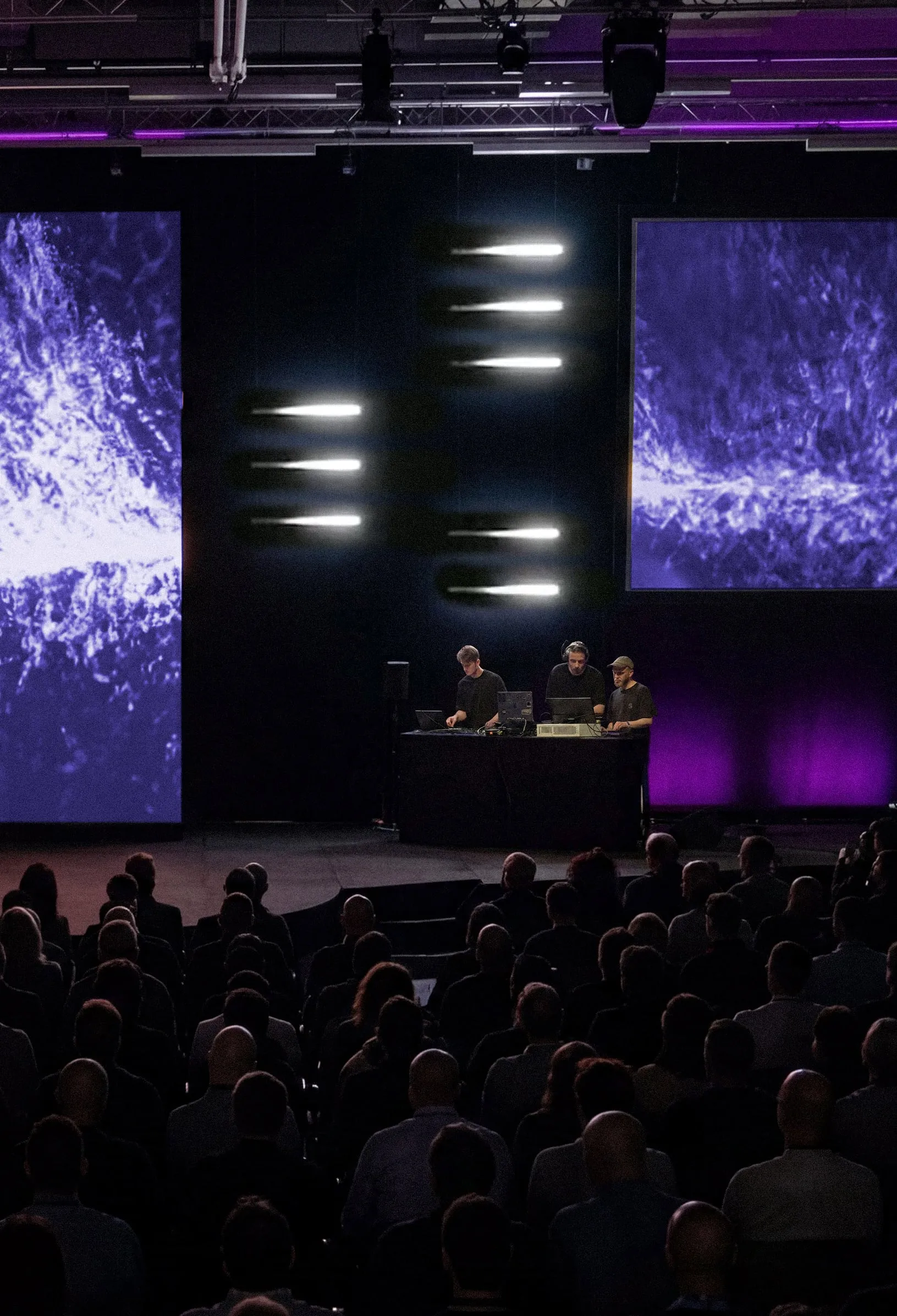 Visuels violets sur les écrans et barres lumineuses au-dessus des performeurs, vus depuis le public